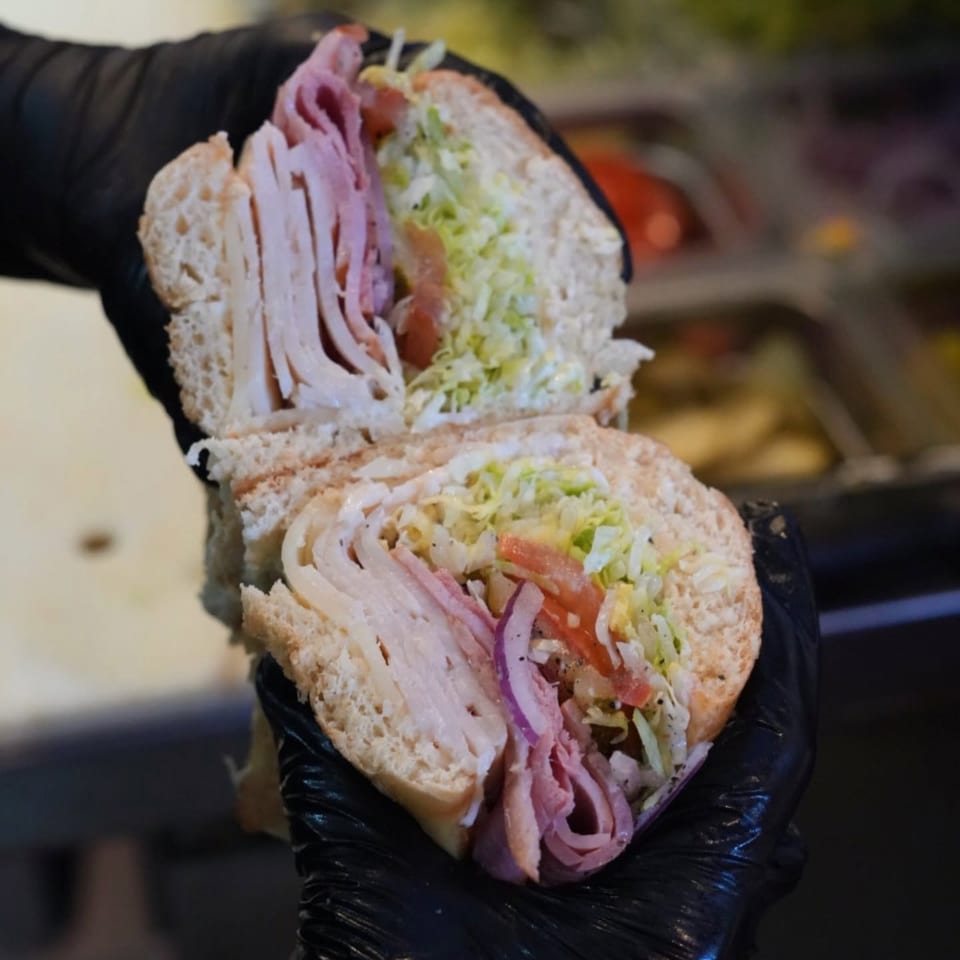 Gloved hands hold a sliced deli sandwich filled with turkey, ham, lettuce, tomato, onion, and dressing.