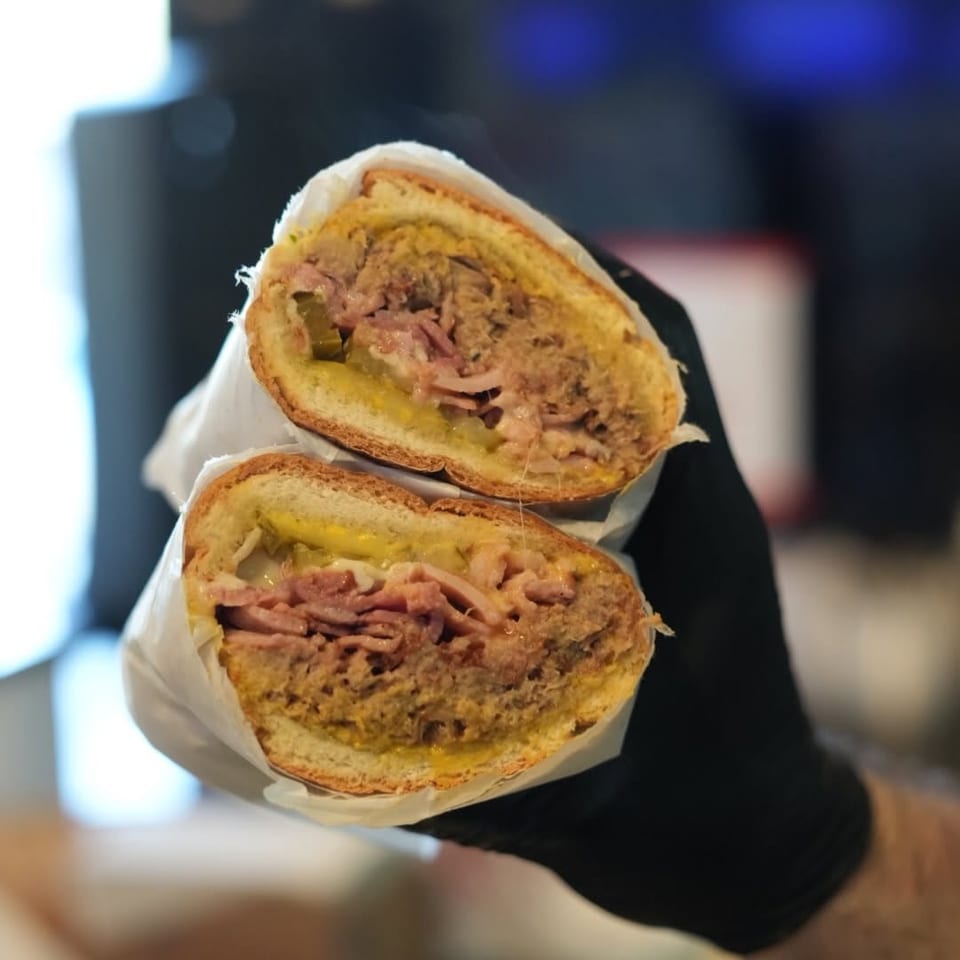 Halved toasted sandwiches reveal layers of meat, cheese, pickles, and sauce, wrapped in paper and held by a gloved hand.