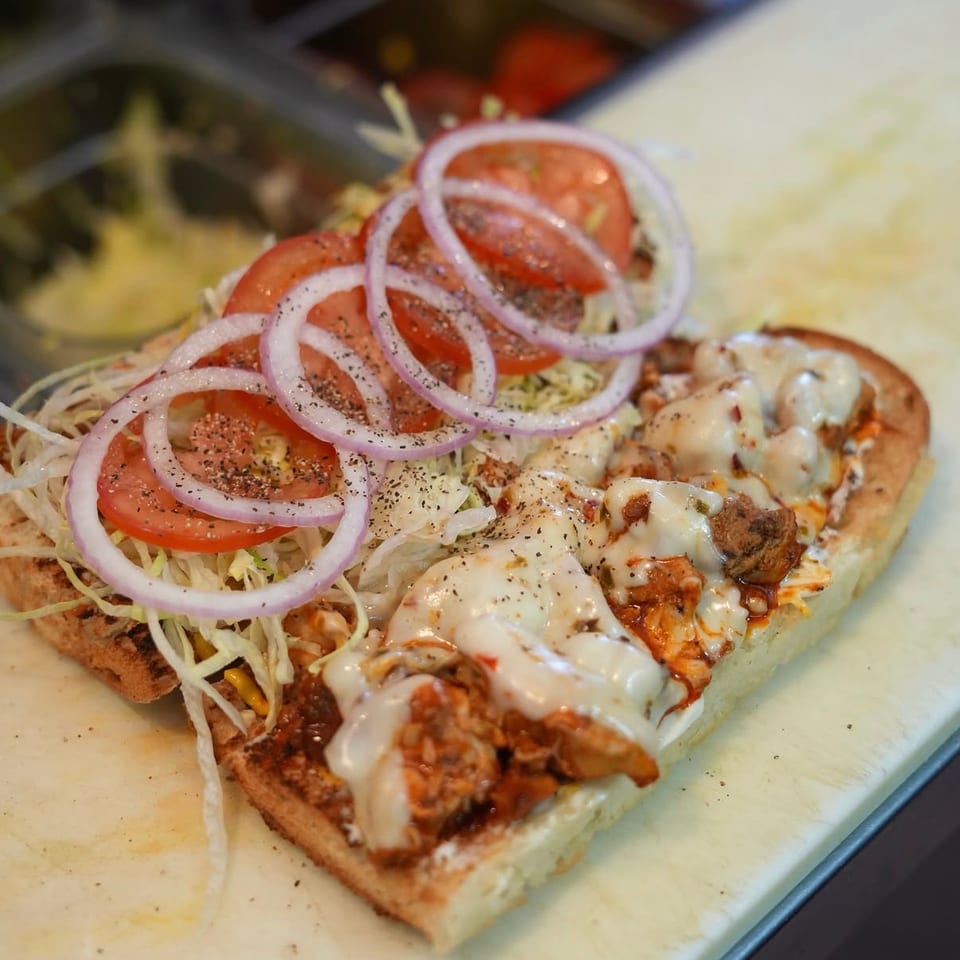 Open sandwich topped with melted cheese, shredded lettuce, tomato slices, and red onion rings on a cutting board.